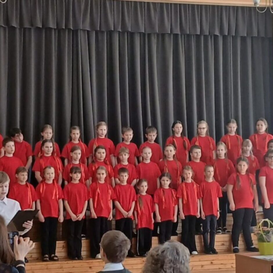 Bērnu koris sarkanos T-kreklos stāv uz skatuves ar tumšu aizkaru fonu. Pa kreisi stāv sieviete blakus klavierēm, bet priekšplānā sēž pieaugušie, skatoties uz uzstāšanos. Zēns korī tur notis.