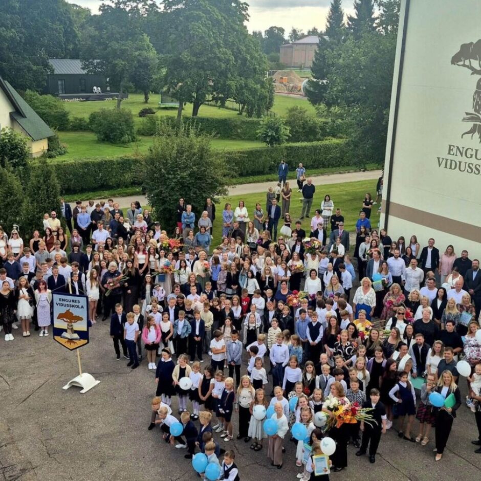 Liela cilvēku grupa, tostarp dažādu vecumu skolēni, skolotāji un vecāki, stāv pie skolas ēkas, uz kuras ir uzraksts 