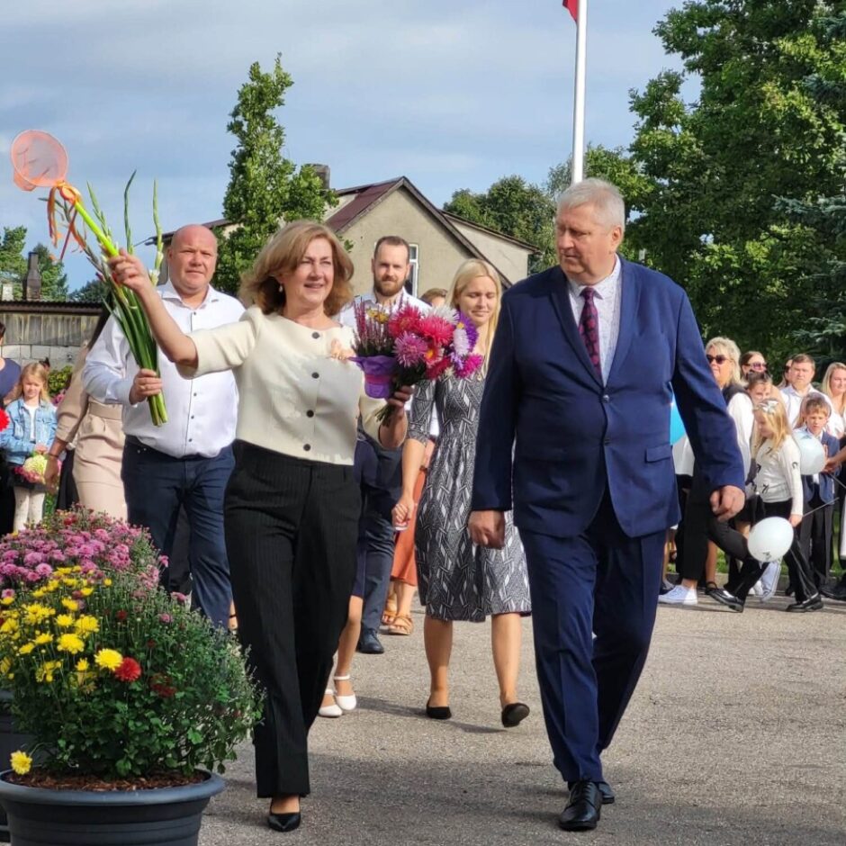 Smaidīga sieviete baltā jaciņā ar ziedu pušķiem rokās soļo blakus vīrietim tumši zilā uzvalkā. Fonā redzama cilvēku grupa, tostarp bērni, un pacelts Latvijas karogs, priekšplānā – ziedu pods ar krāsainām krizantēmām.