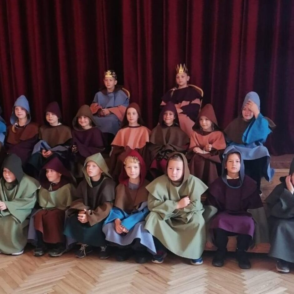 Bērnu grupa, tērpusies krāsainos viduslaiku stila apmetņos ar kapucēm, tostarp divi bērni ar kroņiem. Viņi sēž uz skatuves sarkana aizkara priekšā un pozē fotogrāfijai.