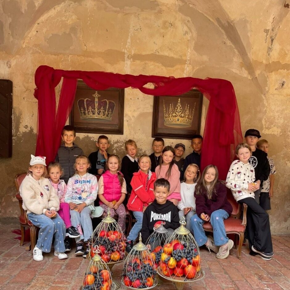 Grupa bērnu, apmēram divdesmit, pozē fotogrāfijai vecas pils telpā ar sarkanām drapērijām un kroņu gleznām pie sienas. Bērni sēž uz krēsliem un stāv, smaidot. Priekšplānā uz grīdas ir vairāki grezni statīvi, kas pildīti ar āboliem, vīnogām un persikiem. Vienai meitenei ir džemperis ar uzrakstu 