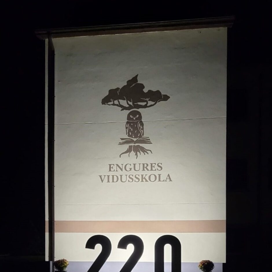 Naktī izgaismota ēkas siena ar Engures vidusskolas logotipu: koka siluets virs pūces, kas sēž uz atvērtas grāmatas. Zem tā ir uzraksts 