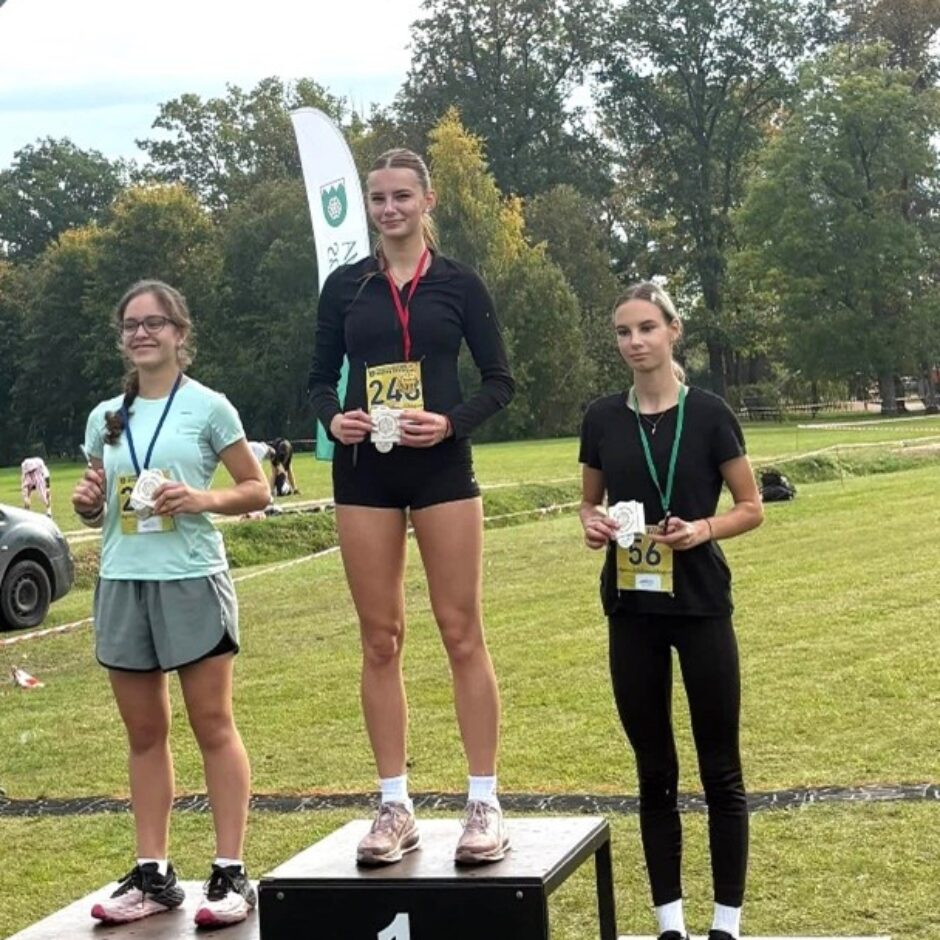 Trīs jaunas sievietes smaida, turot sudraba medaļas uz goda pjedestāla sporta sacensībās. Uz 1. vietas stāv sieviete ar sacensību numuru 243, uz 2. vietas sieviete ar numuru 282, un uz 3. vietas sieviete ar numuru 56. Uz pjedestāla redzams uzraksts 