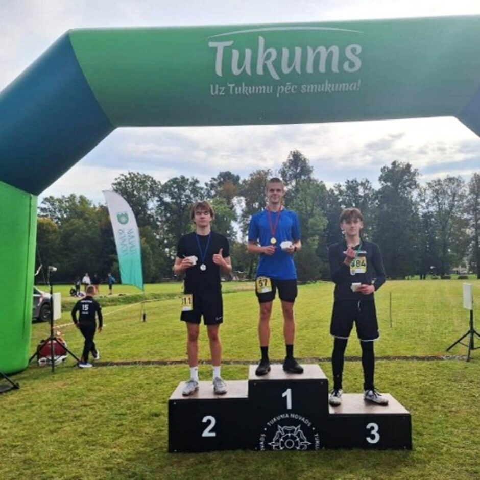 Trīs jauni vīrieši sporta tērpos stāv uz goda pjedestāla ar medaļām un kausiem. Vīrietis 1. vietā ir zilā kreklā, 2. vietā – melnā, rādot 