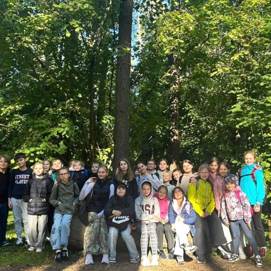Liela bērnu un pusaudžu grupa pozē fotogrāfijai saulainā mežā. Daļa stāv, daļa sēž uz baļķa. Uz viena zēna krekla rakstīts 