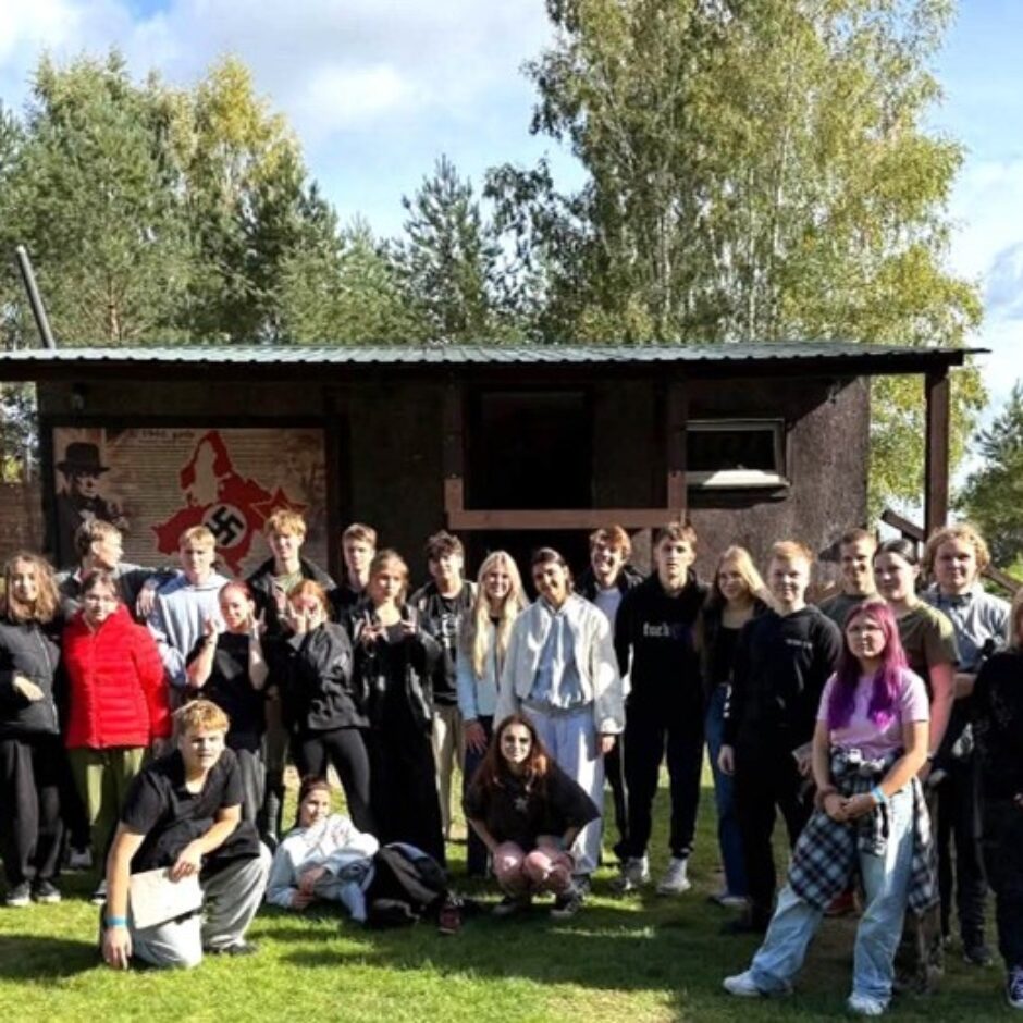Apmēram 30 jauniešu grupa stāv uz zālāja pie tumšas koka ēkas, uz kuras ir plakāts ar nacistiskās Vācijas karti, lielu svastiku, tekstu 