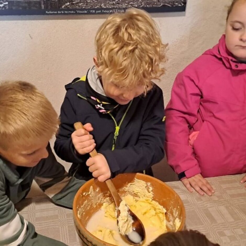 Trīs bērni pie galda. Vidējais zēns ar gaišiem cirtainiem matiem maisa sviestu koka bļodā ar koka karoti. Kreisais zēns un labā meitene vēro. Fonā uz sienas redzams teksts: 