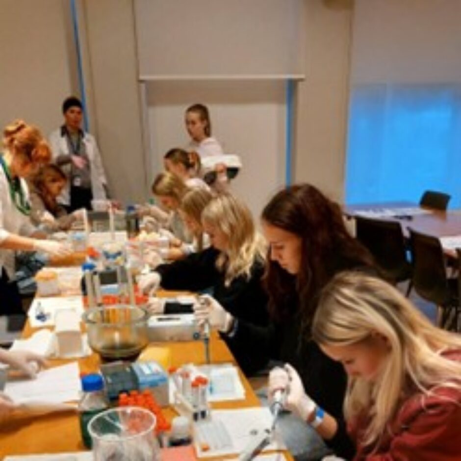Vairāki jaunieši laboratorijas nodarbībā sēž pie galdiem, valkājot cimdus un strādājot ar pipetēm un mēģenēm.