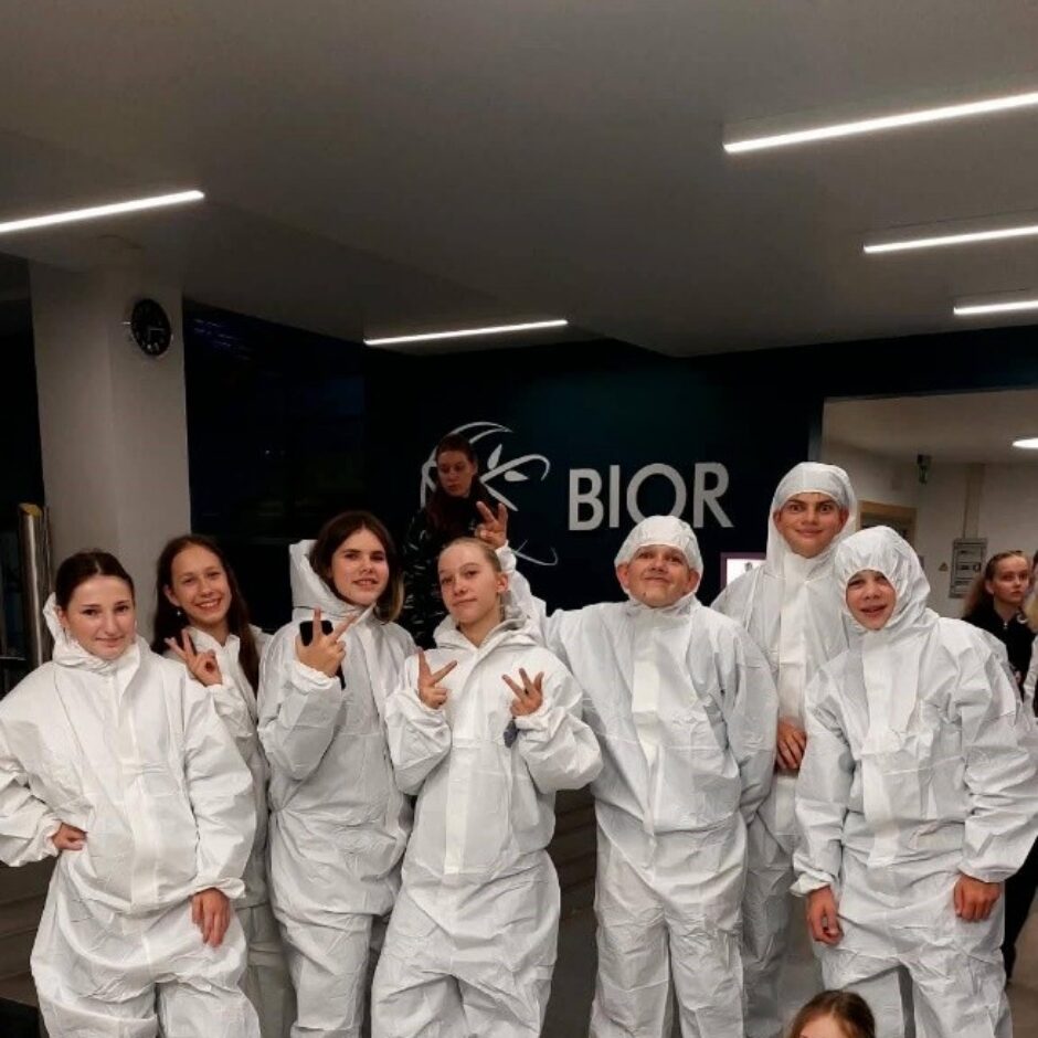 Grupa bērnu un jauniešu baltos aizsargtērpos pozē fotogrāfijai. Septiņi stāv un divi guļ priekšplānā uz grīdas. Daži rāda miera zīmes. Fonā redzams uzraksts 
