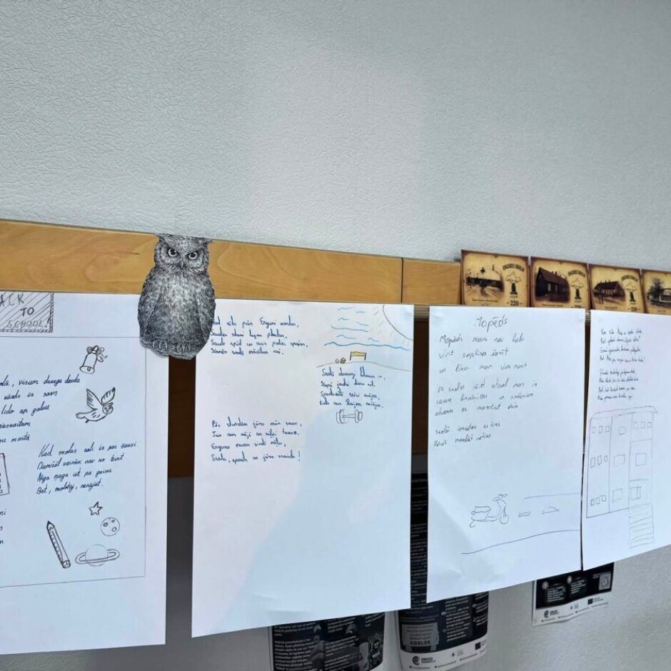 Sienas daļa ar koka plauktu, pie kuras piestiprināti skolēnu darbi un dekori. Kreisajā lapā redzams uzraksts 