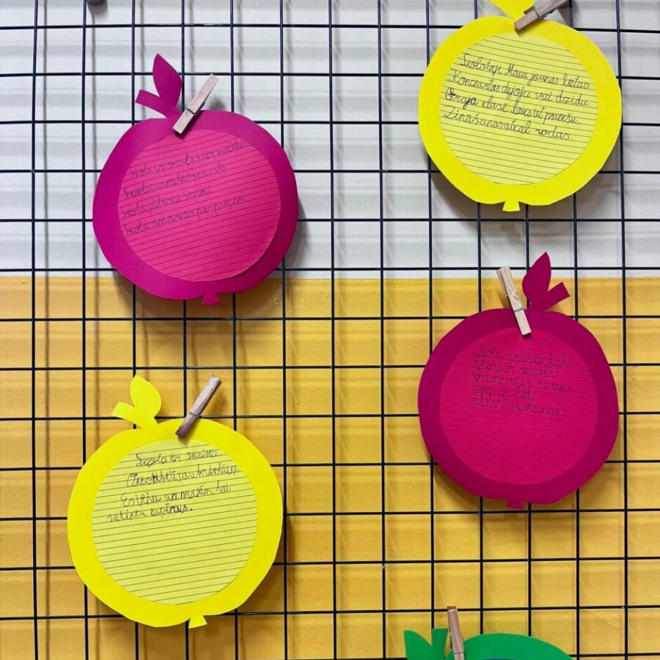 Piecas krāsainas, ābolu formas zīmītes ar rokraksta tekstu ir piestiprinātas pie melnas režģa tāfeles, kas daļēji atklāj dzeltenu fonu. Augšējā kreisajā rozā ābolā rakstīts: 