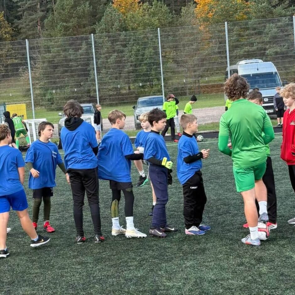 Zēnu grupa zilās un zaļās sporta formās stāv uz mākslīgā seguma futbola laukuma, iespējams, treniņā vai pārtraukumā. Viens zēns spilgti zaļā formā ir vērsts pret grupu zilajos. Fonā redzami zaļi konteineri ar uzrakstiem 