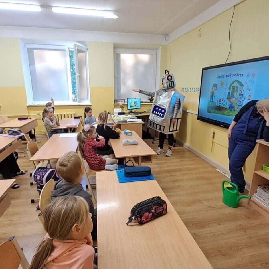 Klasē robota kostīmā tērpta persona ar roku rāda uz lielu ekrānu, kurā redzams teksts 