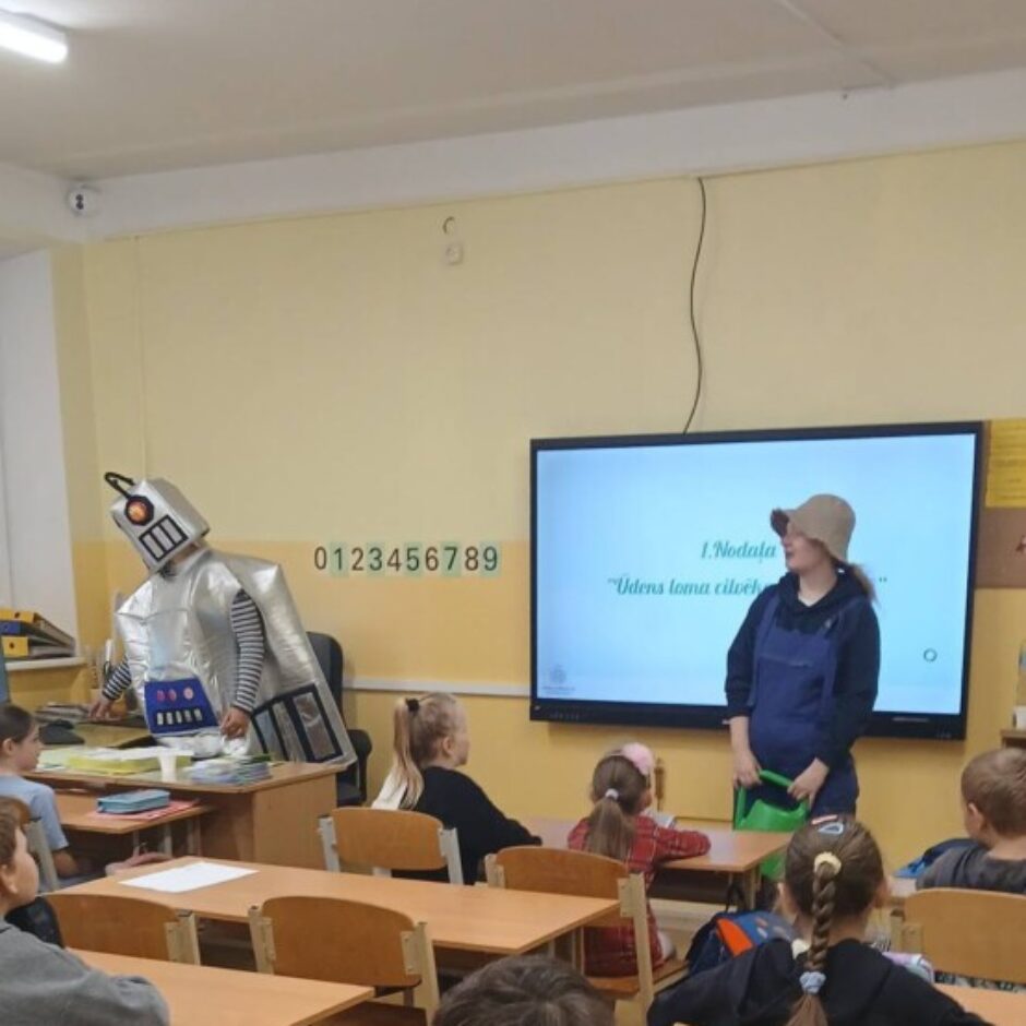 Klasē skolotāja ar cepuri un tumšu kombinezonu stāv pie liela ekrāna, kurā redzams teksts 