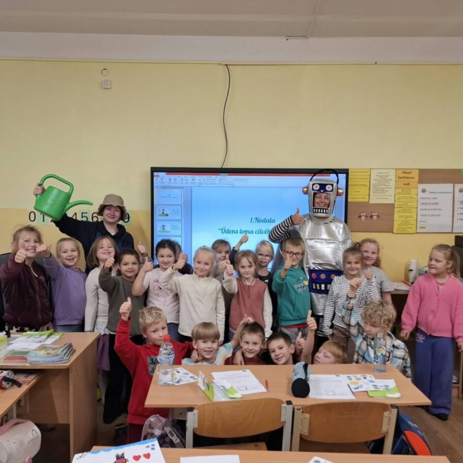 Grupas bērni un divi pieaugušie skolā, lielākā daļa rādot īkšķi uz augšu. Viens pieaugušais ir robota kostīmā, otrs tur zaļu laistāmo kannu. Uz ekrāna fonā redzams teksts: 