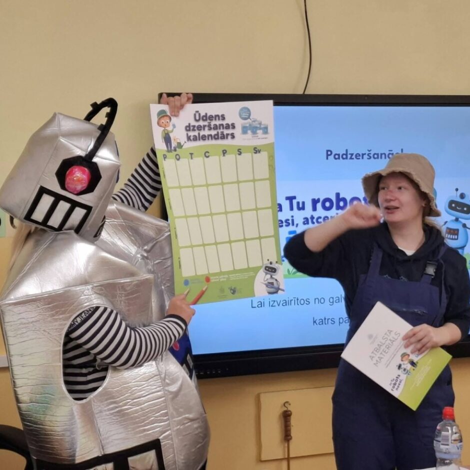 Cilvēks robota kostīmā tur rokās plakātu ar uzrakstu 