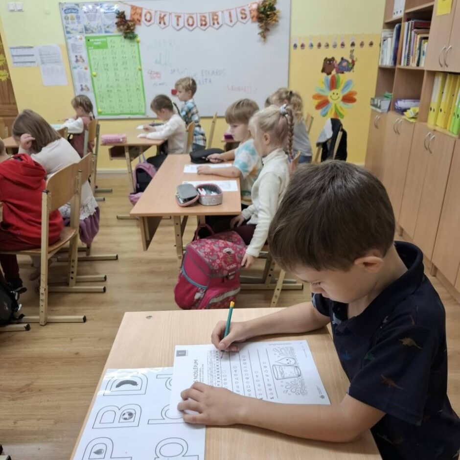 Skolas klasē zēns pirmajā plānā ar zīmuli raksta uz darba lapas, kur redzams daļēji vārds 