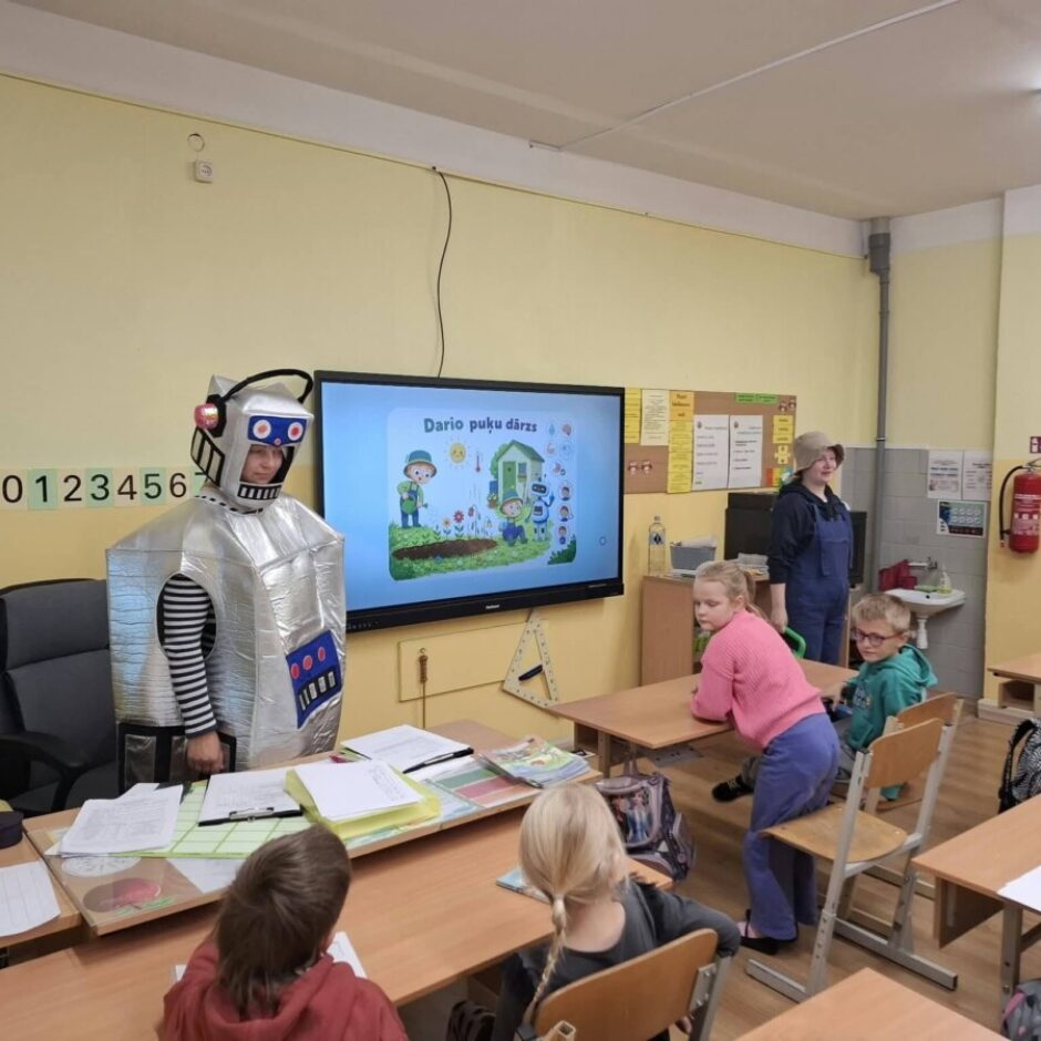 Persona sudraba robota kostīmā stāv klases priekšā pie liela ekrāna, uz kura redzams uzraksts 