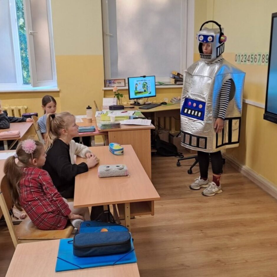 Klasē, kurā bērni sēž pie galdiem, divi pieaugušie rāda priekšnesumu; viens ir sudrabainā robota kostīmā, bet otrs, ģērbies kā dārznieks ar platmali un kombinezonu, tur plastmasas pudeli un stāv pie ekrāna, uz kura redzams uzraksts “Dario puķu dārzs” un zīmējums ar dārzu.