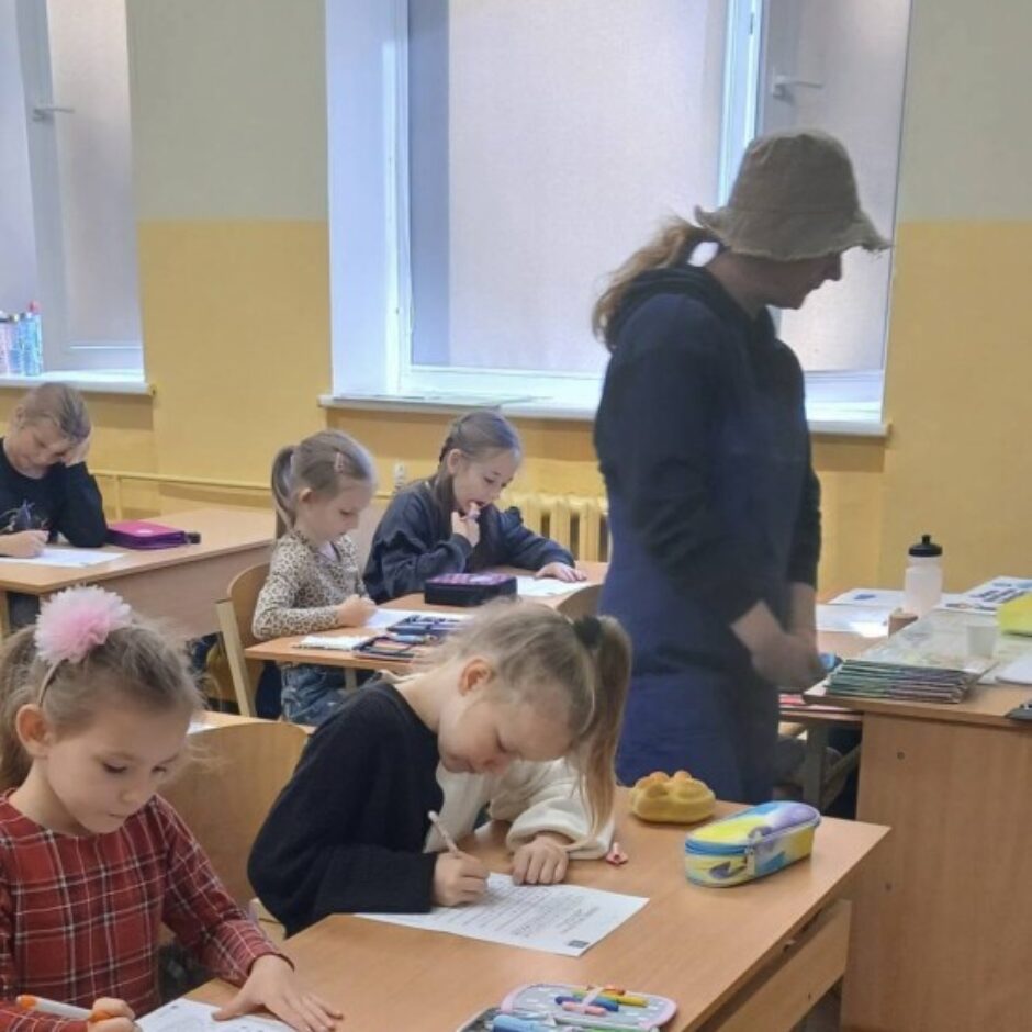Bērni klasē sēž pie galdiem un raksta vai zīmē uz papīriem. Fonā stāv pieauguša sieviete cepurē, uzraugot. Uz galdiem redzamas zīmuļu kārbas ar krāsainiem zīmuļiem, bet pie viena galda karājas zila mugursoma.