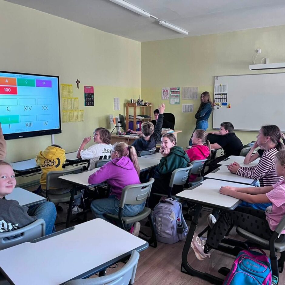 Gaišā klasē bērni piedalās mācību stundā. Priekšpusē redzams liels interaktīvs ekrāns ar skaitļiem un romiešu cipariem: 20, 4, 9, 14, 13, 100, IX, C, XIV, XX, VI, I, XIII, IV. Vairākiem skolēniem ir paceltas rokas, un sieviete skolotāja stāv pie tāfeles. Meitene priekšplānā smaida kamerā.
