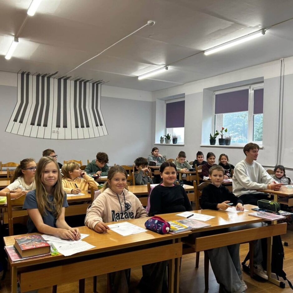 Mūzikas klase ar vairākiem bērniem un pusaudžiem, kas sēž pie koka galdiem. Daži skolēni smaida un skatās kamerā. Aizmugurē uz sienas ir liels, stilizēts klavieru taustiņu zīmējums. Vienai meitenei ir džemperis ar uzrakstu 