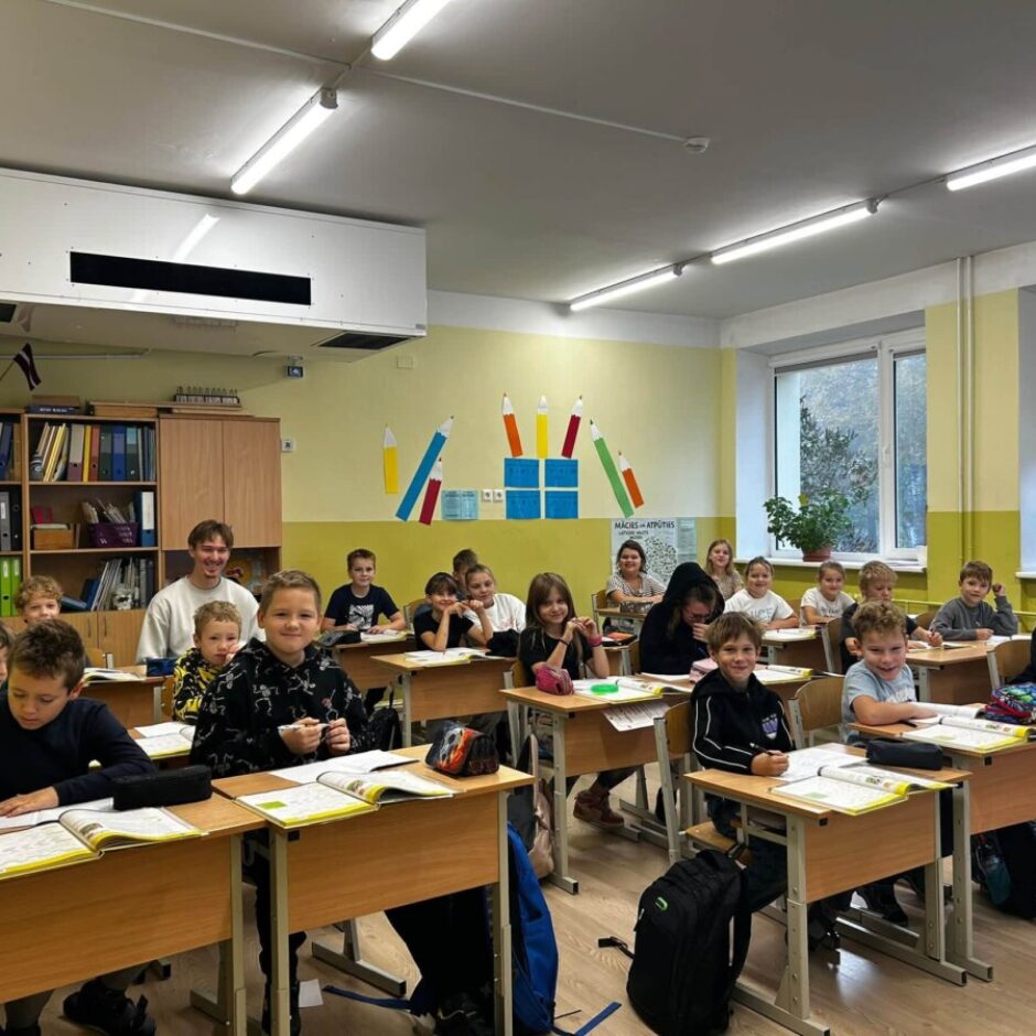 Smaidīgi skolēni un vīrietis sēž gaišā klasē. Daži bērni raksta, citi skatās kamerā. Uz sienas ir plakāts ar uzrakstu 