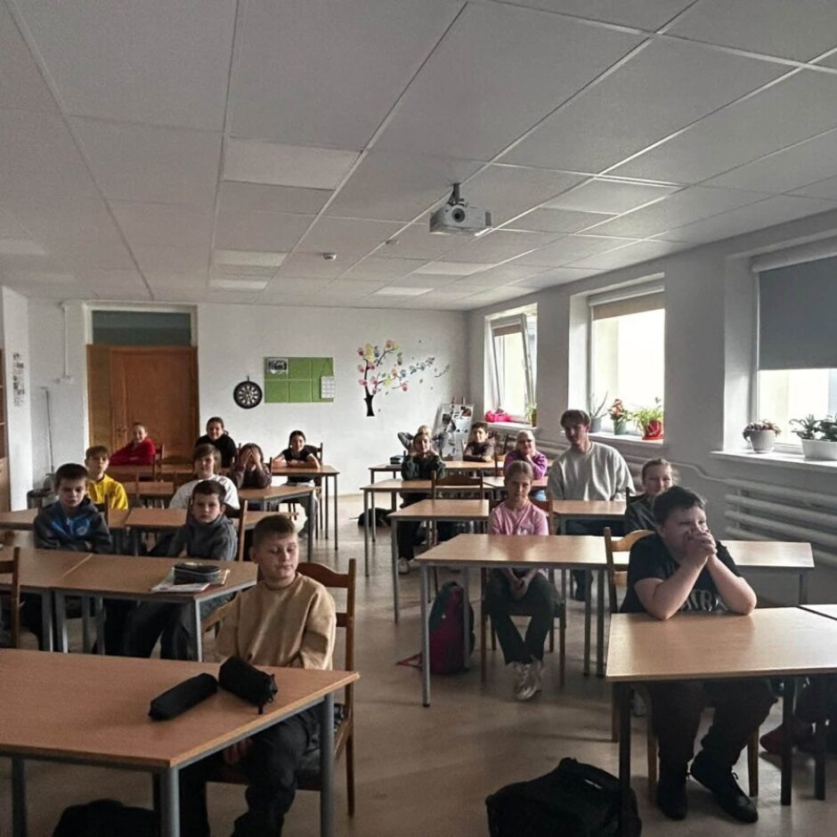 Skolēnu grupa sēž gaišā klasē pie koka galdiem, daži skatās uz priekšu. Telpā ir logi ar žalūzijām un puķupodiem, kā arī griestu projektors.
