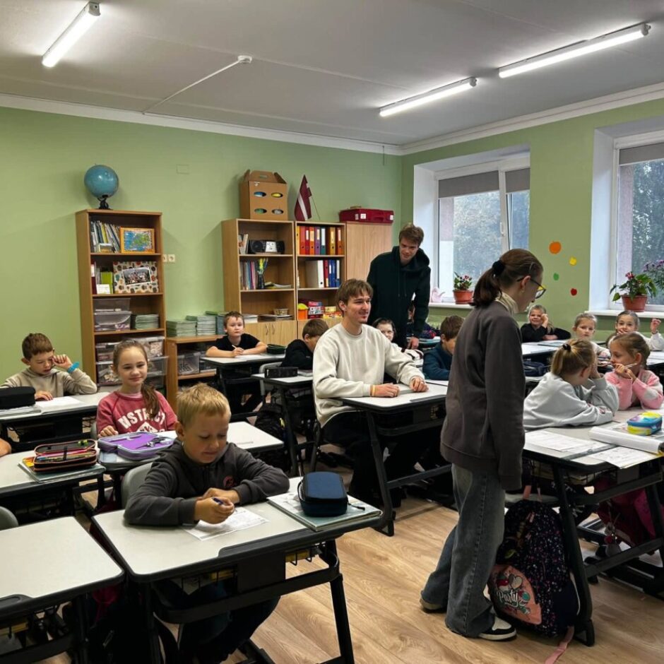 Plašā klases telpā pie galdiem sēž apmēram piecpadsmit skolēni, daži skatās kamerā un smaida, citi ir iegrimuši uzdevumos. Divi pieaugušie stāv starp galdiem, viens no tiem sarunājas ar skolēniem. Fonā redzami grāmatu plaukti, globuss un Latvijas karogs.