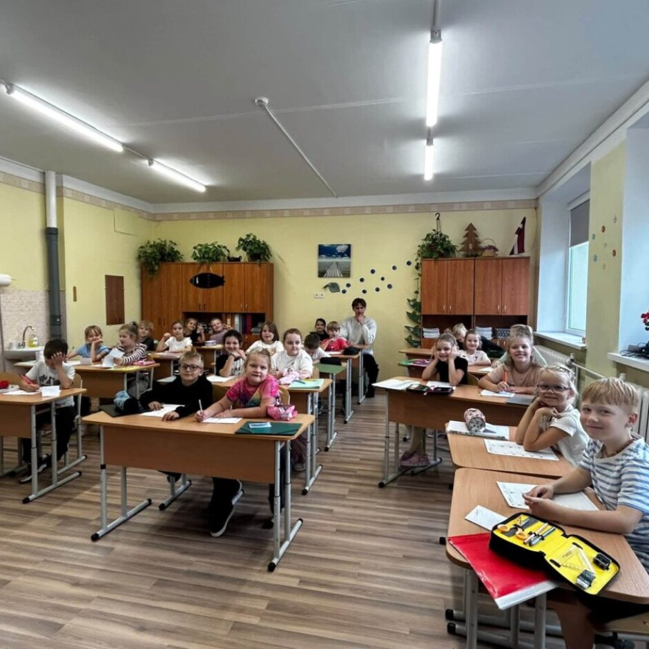 Gaišā klasē sākumskolas vecuma bērni sēž pie koka galdiem. Vairāki bērni smaida, skatoties uz kameru, kamēr citi nodarbojas ar rakstīšanu. Fonā pie galda sēž pieaugušais (skolotājs), bet uz palodzes redzami ziedi.