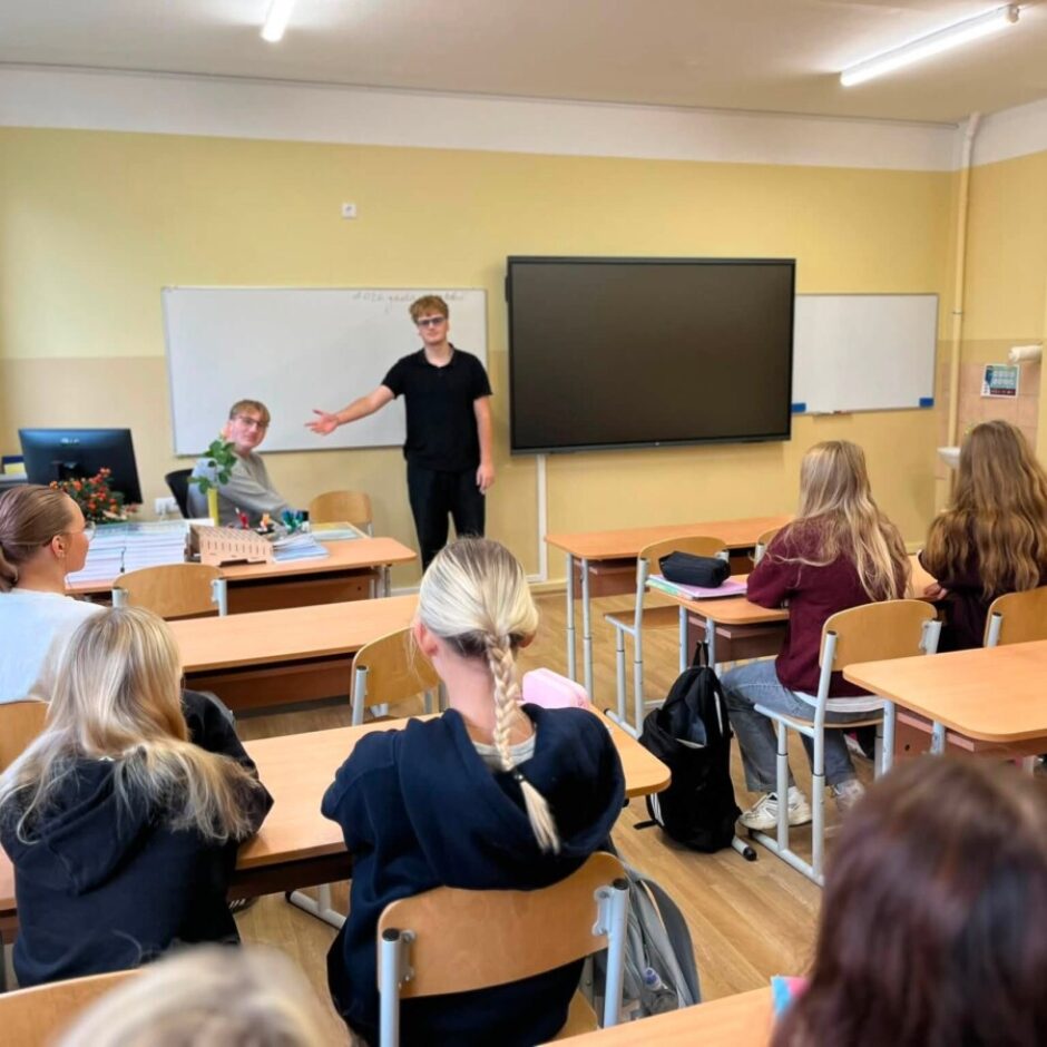 Klasē jauns vīrietis melnā T-kreklā stāv pie tāfeles, žestikulējot uz jaunu vīrieti pelēkā džemperī, kurš sēž pie galda. Vairākas jaunas sievietes sēž pie galdiem, pagriezušās ar mugurām. Aizmugurē redzama balta tāfele un liels melns ekrāns.