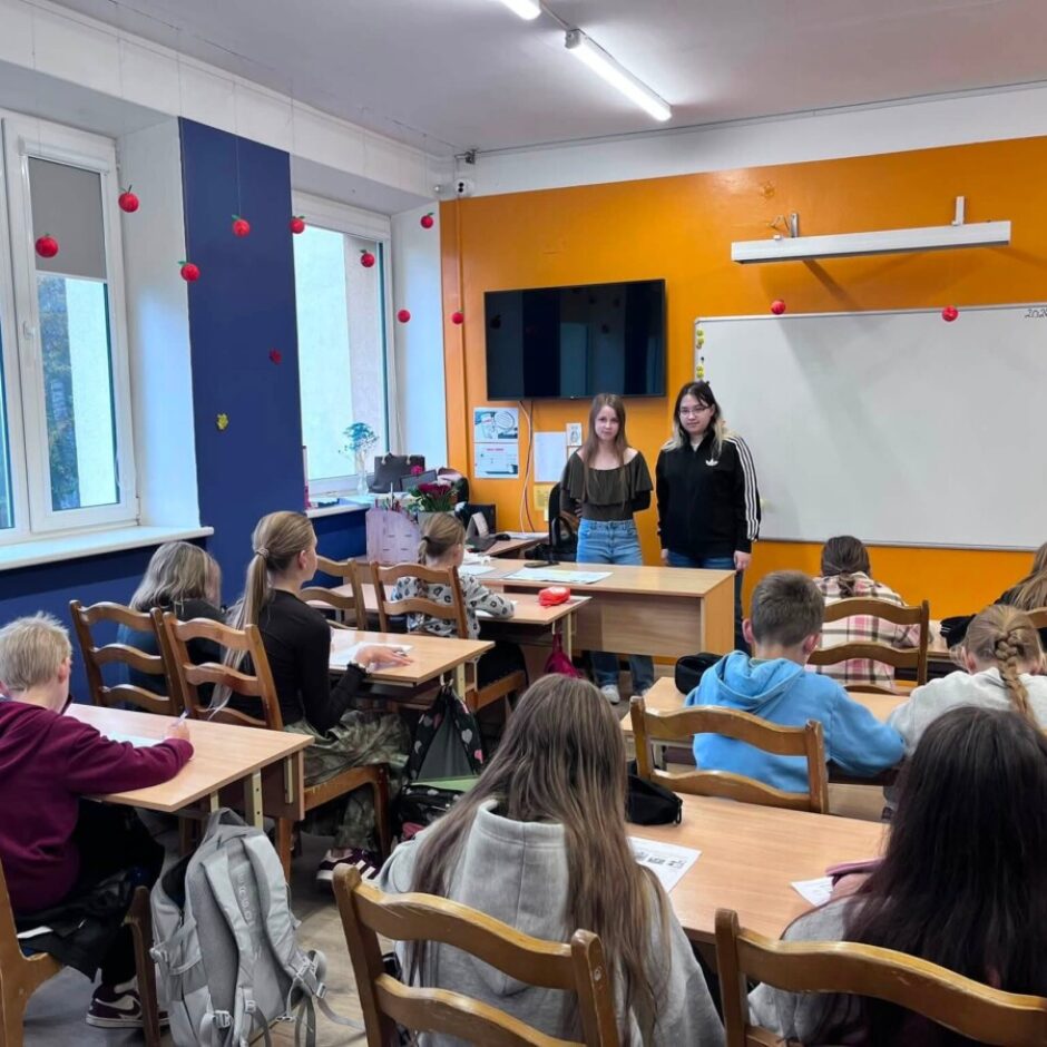 Skolēnu klase, kurā divas jaunietes stāv pie apelsīnu krāsas sienas, pie baltas tāfeles ar uzrakstu 