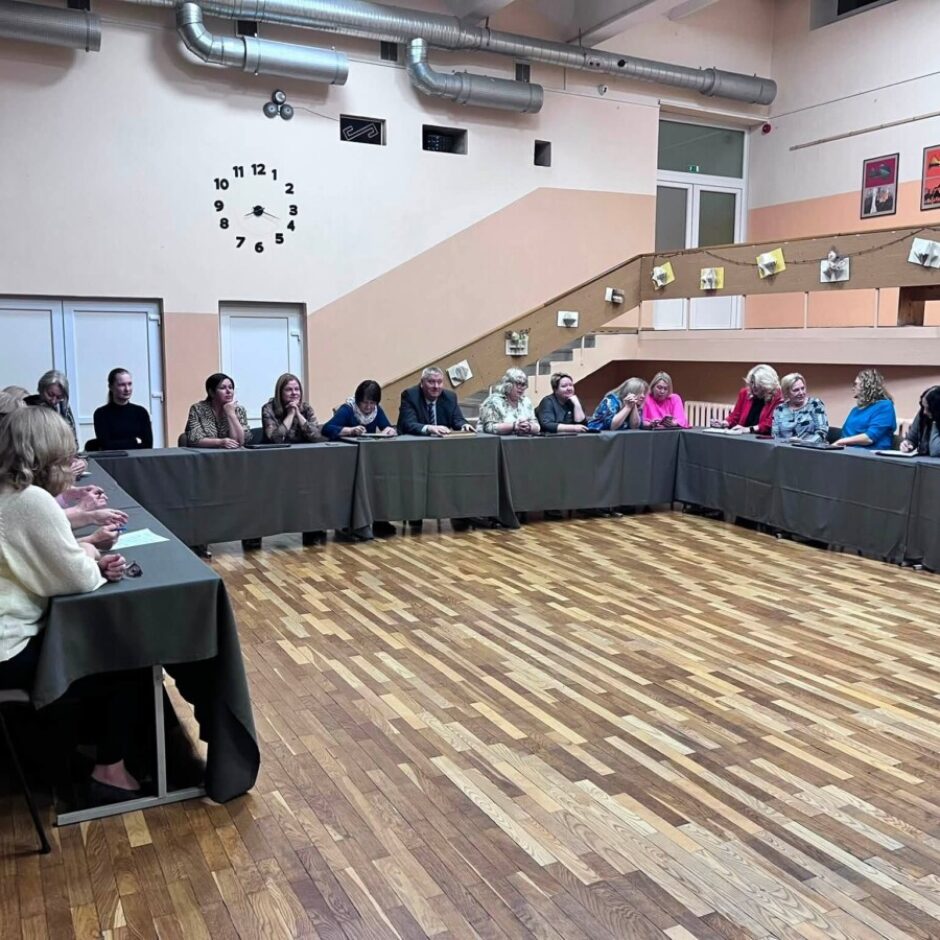 Cilvēku grupa sēž pie U veida galda sanāksmē telpā ar koka grīdu. Sienas rotā liels pulkstenis un dažādi mākslas darbi.