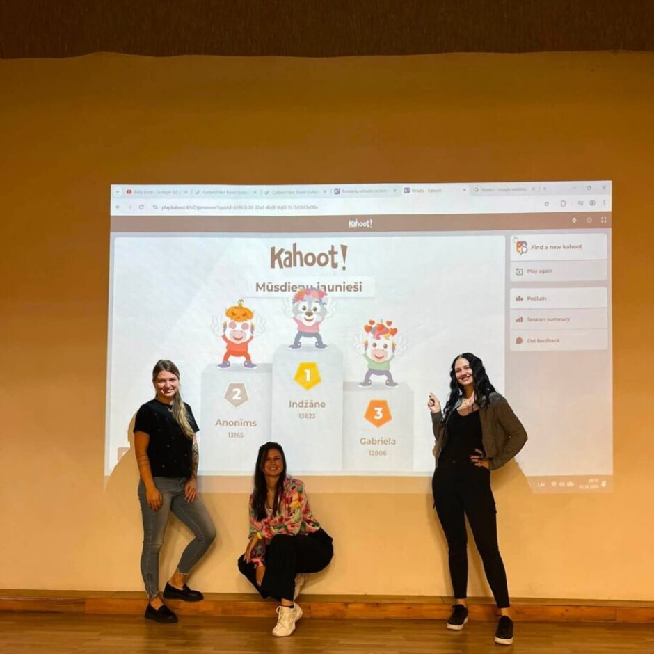 Trīs jaunas sievietes pozē pie liela projektora ekrāna, kurā redzami Kahoot! spēles rezultāti. Ekrānā ir uzraksti 