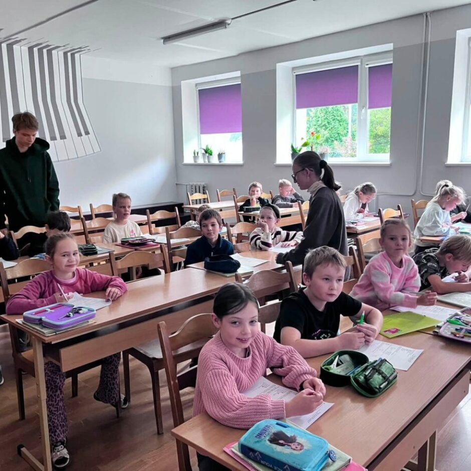 Klašu telpa ar aptuveni divdesmit jauniem skolēniem, kas sēž pie koka galdiem. Pieaugušais vīrietis tumšā džemperī stāv kājās starp galdiem, uzraugot bērnus, no kuriem daži raksta, bet citi skatās uz priekšu. Sienu rotā liels klavieru taustiņu sienas zīmējums. Priekšplānā meitene rozā džemperī smaida kamerā.