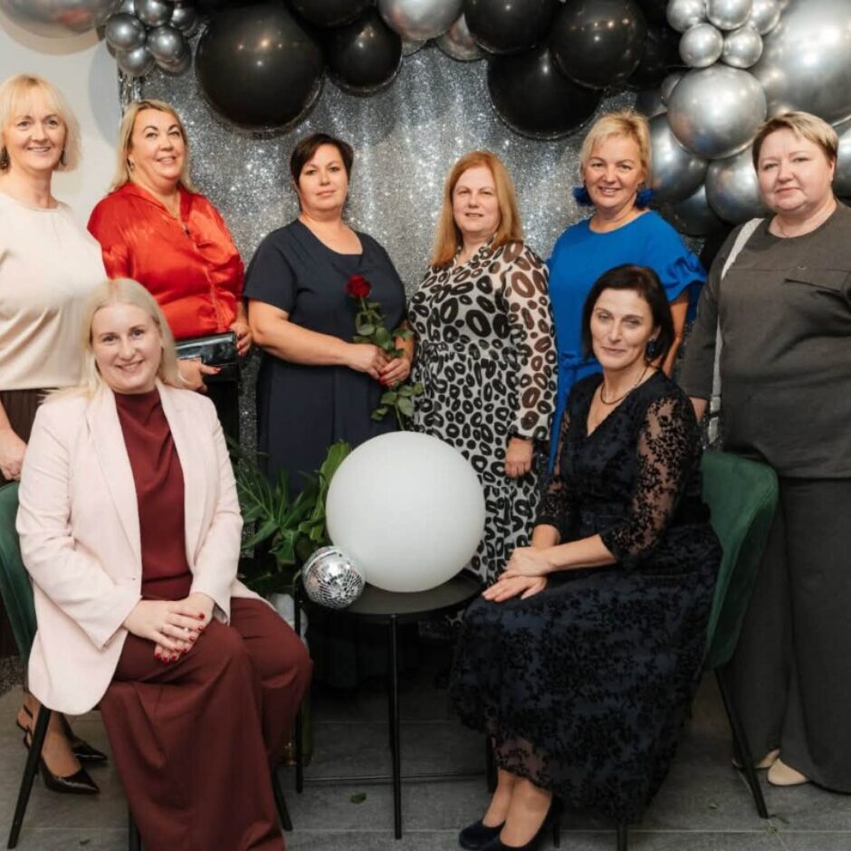 Astoņas smaidīgas sievietes pozē grupveida fotoattēlā pasākumā. Sešas sievietes stāv, bet divas sēž priekšplānā. Fonā redzami melni un sudrabaini baloni. Viena sieviete tur sarkanu rozi.