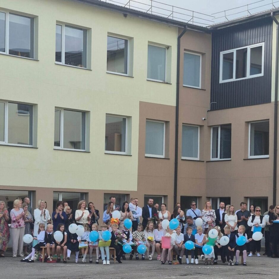 Skolas pagalmā svinīgā pasākumā bērni, iespējams, pirmklasnieki, sēž uz soliem ar ziliem un baltiem baloniem un ziediem, bet pieaugušie aizmugurē aplaudē pie gaišas skolas ēkas.