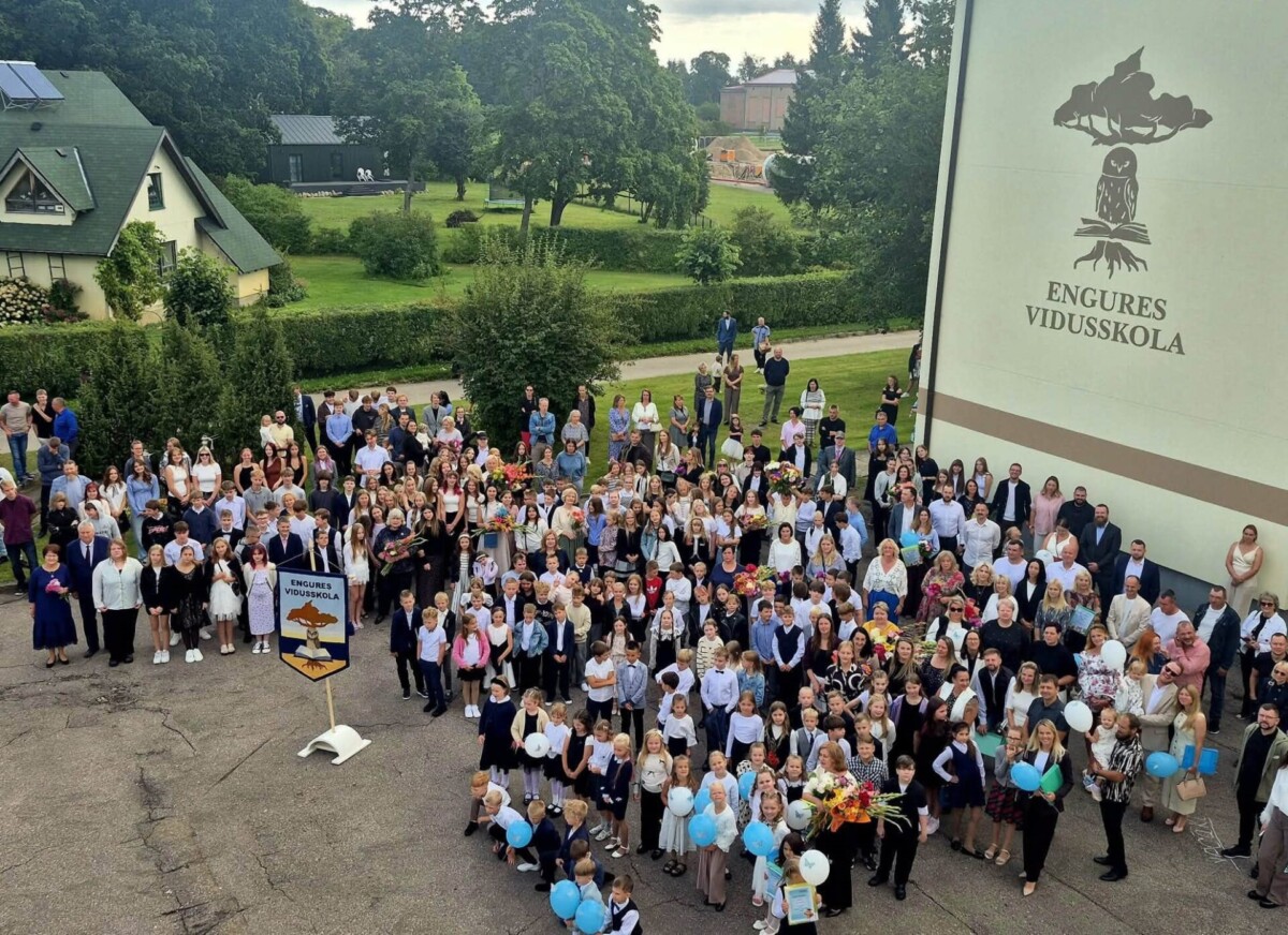 Liela cilvēku grupa, tostarp dažādu vecumu skolēni, skolotāji un vecāki, stāv pie skolas ēkas, uz kuras ir uzraksts 