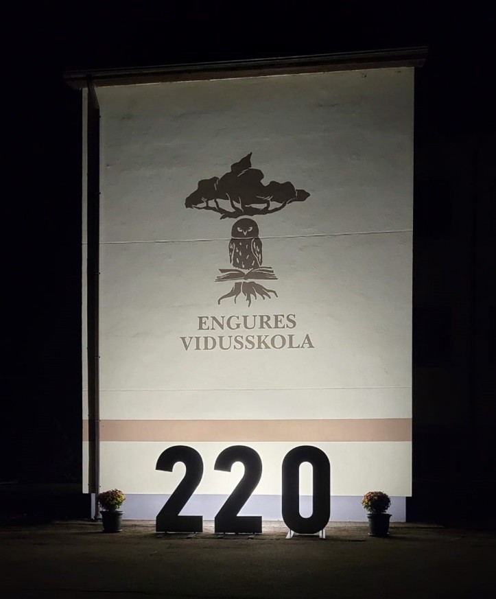 Naktī izgaismota ēkas siena ar Engures vidusskolas logotipu: koka siluets virs pūces, kas sēž uz atvērtas grāmatas. Zem tā ir uzraksts 