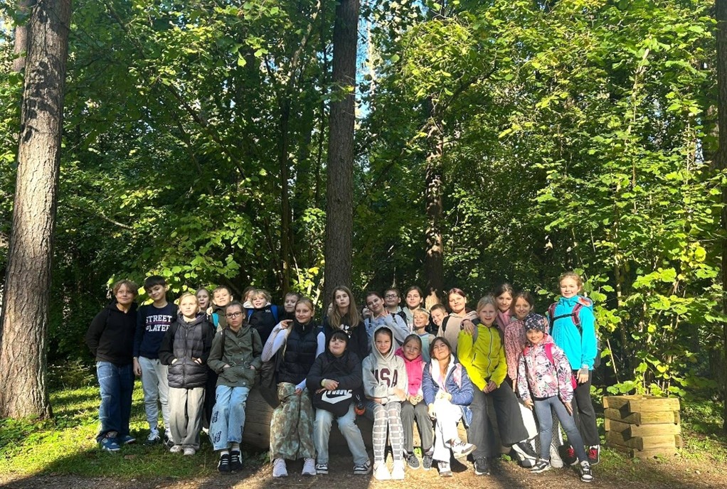 Liela bērnu un pusaudžu grupa pozē fotogrāfijai saulainā mežā. Daļa stāv, daļa sēž uz baļķa. Uz viena zēna krekla rakstīts 