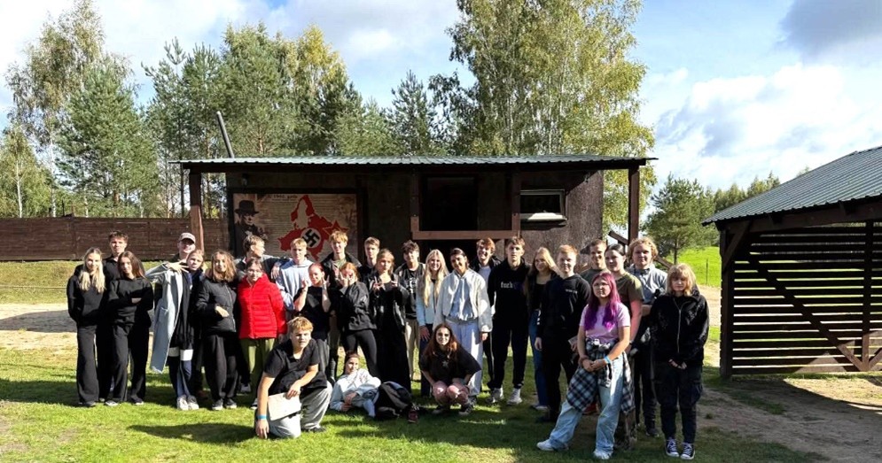 Apmēram 30 jauniešu grupa stāv uz zālāja pie tumšas koka ēkas, uz kuras ir plakāts ar nacistiskās Vācijas karti, lielu svastiku, tekstu 