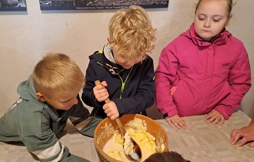 Trīs bērni pie galda. Vidējais zēns ar gaišiem cirtainiem matiem maisa sviestu koka bļodā ar koka karoti. Kreisais zēns un labā meitene vēro. Fonā uz sienas redzams teksts: 