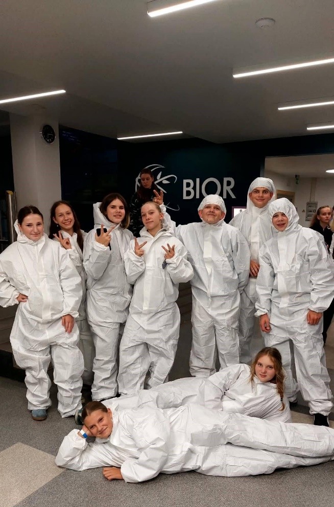 Grupa bērnu un jauniešu baltos aizsargtērpos pozē fotogrāfijai. Septiņi stāv un divi guļ priekšplānā uz grīdas. Daži rāda miera zīmes. Fonā redzams uzraksts 