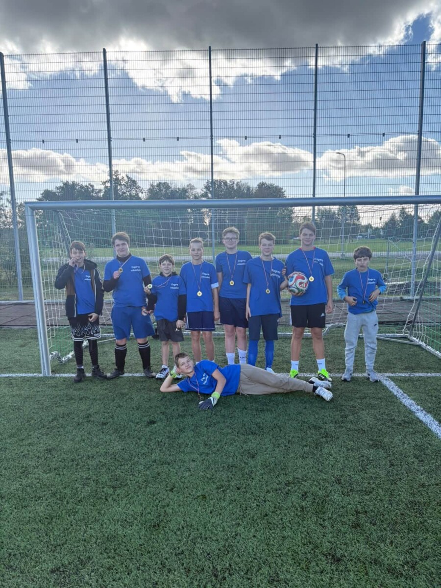 Astoņi zēni futbola laukumā, septiņi stāvot un viens guļot priekšplānā, pozē fotogrāfijai vārtu priekšā. Viņi valkā zilus kreklus ar uzrakstu 