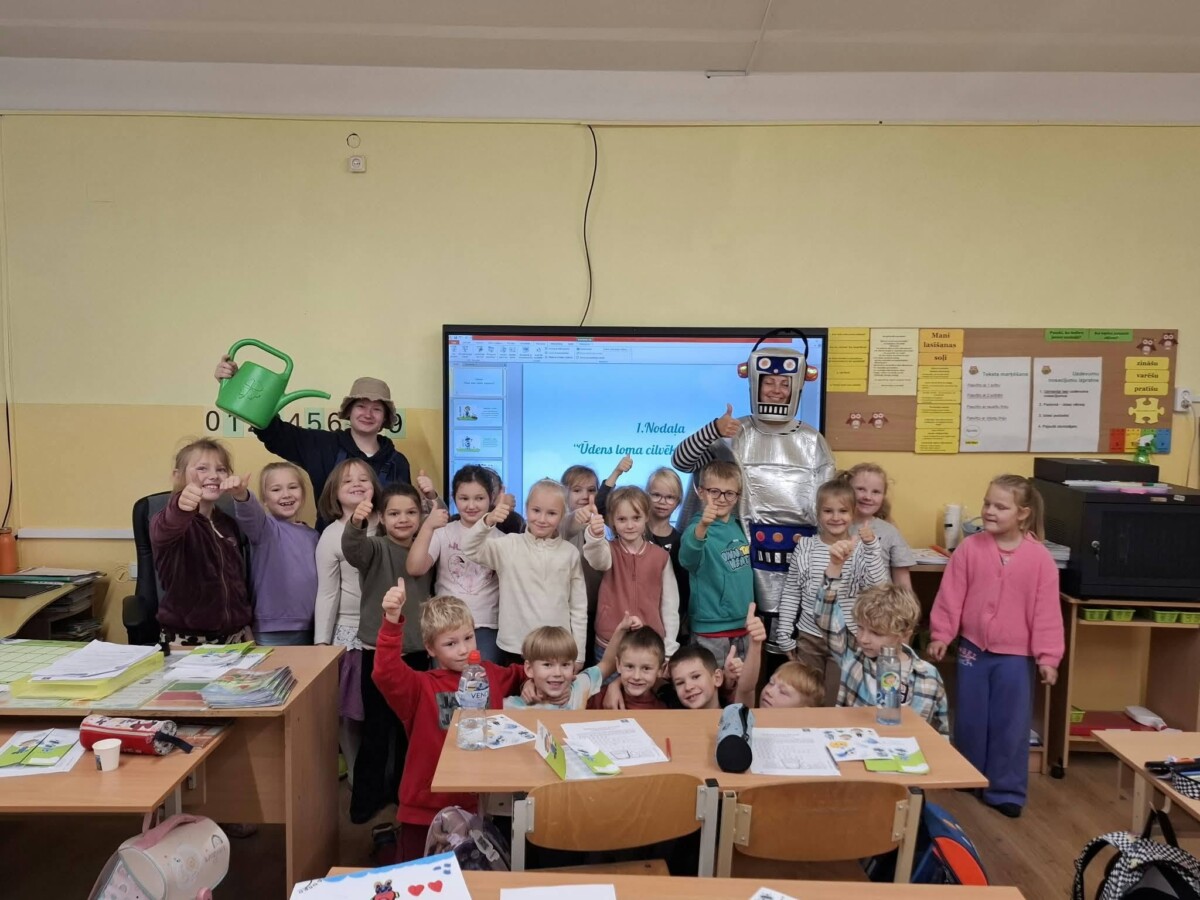 Grupas bērni un divi pieaugušie skolā, lielākā daļa rādot īkšķi uz augšu. Viens pieaugušais ir robota kostīmā, otrs tur zaļu laistāmo kannu. Uz ekrāna fonā redzams teksts: 