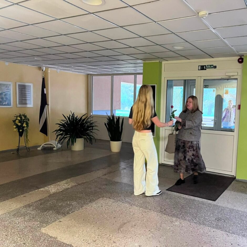 Divas sievietes skolas gaiteņa durvju priekšā apmainās ar sarkanu rozi. Jaunāka sieviete, redzama no muguras, pasniedz rozi vecākai sievietei. Virs durvīm ir zīme 