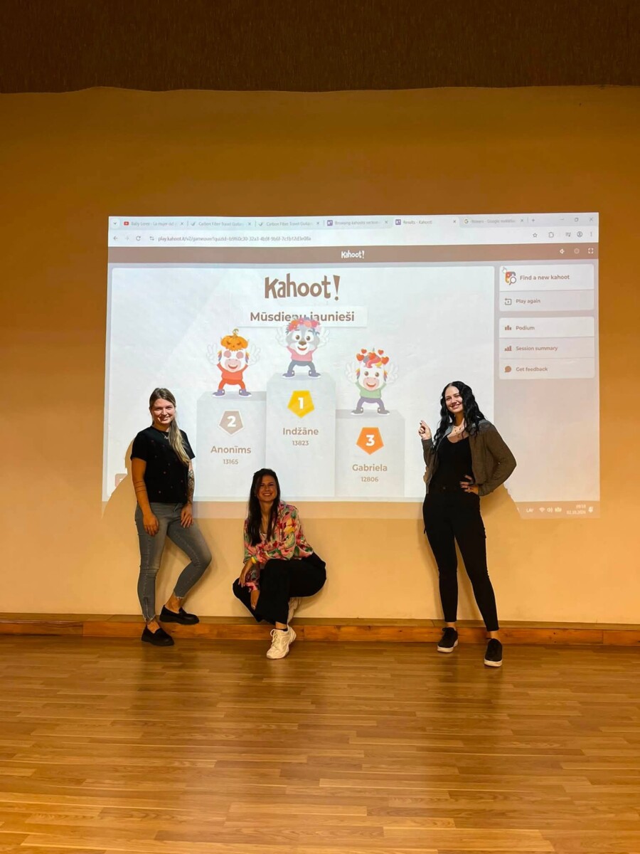 Trīs jaunas sievietes pozē pie liela projektora ekrāna, kurā redzami Kahoot! spēles rezultāti. Ekrānā ir uzraksti 
